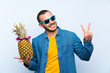 © luismolinero - Colombian man holding a pineapple with sunglasses smiling and showing victory sign
