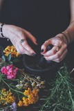 Fototapeta Lawenda - Female wiccan witch grinding summer herbs and flowers with pestle and mortar. Colorful fresh flowers and rosemary on a black table. A woman wearing vintage jewelry holding pestle in her hand. Vertical