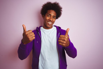 Young african american man wearing purple sweatshirt standing over isolated pink background success sign doing positive gesture with hand, thumbs up smiling and happy. Cheerful expression 