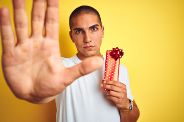 Young caucasian man holding birthday present over yellow isolated background with open hand doing stop sign with serious and confident expression, defense gesture