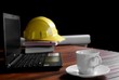 © Prapat - Focus on white ceramic coffee cup with blurred black laptop and yellow safety hard hat and blueprint on engineer office desk in black background