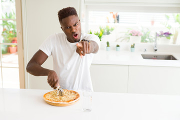 Sticker - African american man eating cheese pizza at home pointing with finger to the camera and to you, hand sign, positive and confident gesture from the front