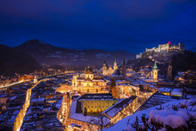 Elevated View Of Salzburg City By Night On A Winter Evening With The Roofs Covered With Snow And The Hohensalzburg Castle