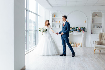 Wall Mural - Beautiful bride in white dress and groom in suit, posing in white Studio interior, wedding