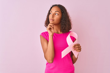 Wall Mural - Young brazilian woman holding cancer ribbon standing over isolated pink background serious face thinking about question, very confused idea