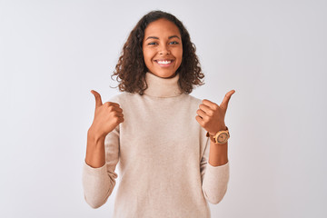 Young brazilian woman wearing turtleneck sweater standing over isolated white background success sign doing positive gesture with hand, thumbs up smiling and happy. Cheerful expression 