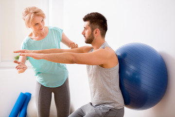 Canvas Print - Young man with back injury exercising with blue gymnastic ball during appointment with female physiotherapist