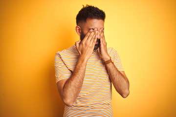 Young indian man wearing t-shirt standing over isolated yellow background rubbing eyes for fatigue and headache, sleepy and tired expression. Vision problem