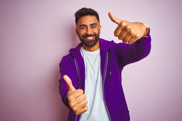 Sticker - Young indian man wearing purple sweatshirt standing over isolated pink background approving doing positive gesture with hand, thumbs up smiling and happy for success. Winner gesture.