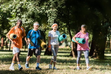 Wall Mural - cheerful senior and multicultural pensioners holding fitness mats and walking in park