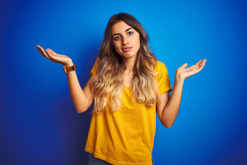 Canvas Print - Young beautiful woman wearing yellow t-shirt over blue isolated background clueless and confused expression with arms and hands raised. Doubt concept.