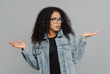 Confused Unaware Afro Woman With Crisp Hair, Raises Hands In Bewilderment, Looks Aside, Cannot Make Decision, Wears Spectacles And Jean Jacket, Isolated Over Grey Background. What Should I Do Now?