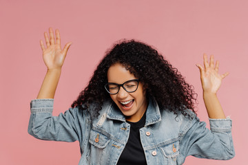 Wall Mural - Isolated shot of beautiful happy woman with curly hairstyle, keeps hands raised, laughs from positive emotions, celebrates success has fun with friends dressed in fashionable apparel. Emotions concept