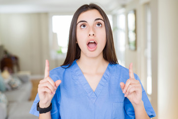 Poster - Beautiful young nurse woman at the clinic amazed and surprised looking up and pointing with fingers and raised arms.