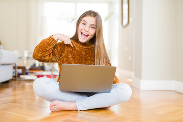 Beautiful young girl studying using laptop sitting on the floor at home with surprise face pointing finger to himself