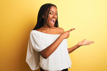 Young african american woman wearing white t-shirt standing over isolated yellow background amazed and smiling to the camera while presenting with hand and pointing with finger.