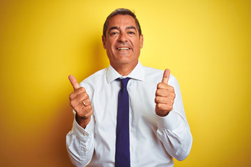 Handsome middle age businessman standing over isolated yellow background success sign doing positive gesture with hand, thumbs up smiling and happy. Cheerful expression and winner gesture.