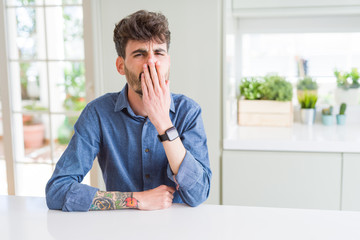 Poster - Young man wearing casual shirt sitting on white table bored yawning tired covering mouth with hand. Restless and sleepiness.