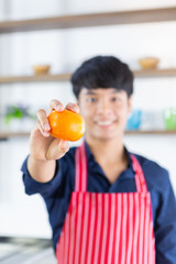 Young smiling asian man with red apron showing orange in right hand on wooden shelf background of kitchen room