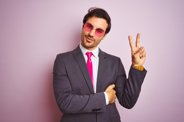 Sticker - Young handsome businessman wearing suit and sunglasses over isolated pink background smiling with happy face winking at the camera doing victory sign. Number two.