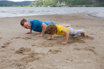  Father and son spend time together and lead a healthy lifestyle outdoors. Man and boy doing push ups are working out. Father and son are doing exercise on the beach in the summer.