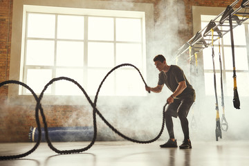 CrossFit training. Side view of young athletic man with perfect body doing crossfit exercises with a rope in the gym.