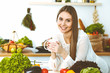 © rogerphoto - Young happy woman is holding white cup and looking at the camera while sitting at wooden table in the kitchen among green vegetables