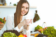 © rogerphoto - Young happy woman is holding white cup and looking at the camera while sitting at wooden table in the kitchen among green vegetables