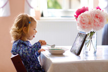 Adorable Toddler Girl Eating Healthy Cereal With Milk For Breakfast And Watching Cartoons On Tablet Pc. Cute Happy Baby Child In Colorful Clothes Sitting In Kitchen