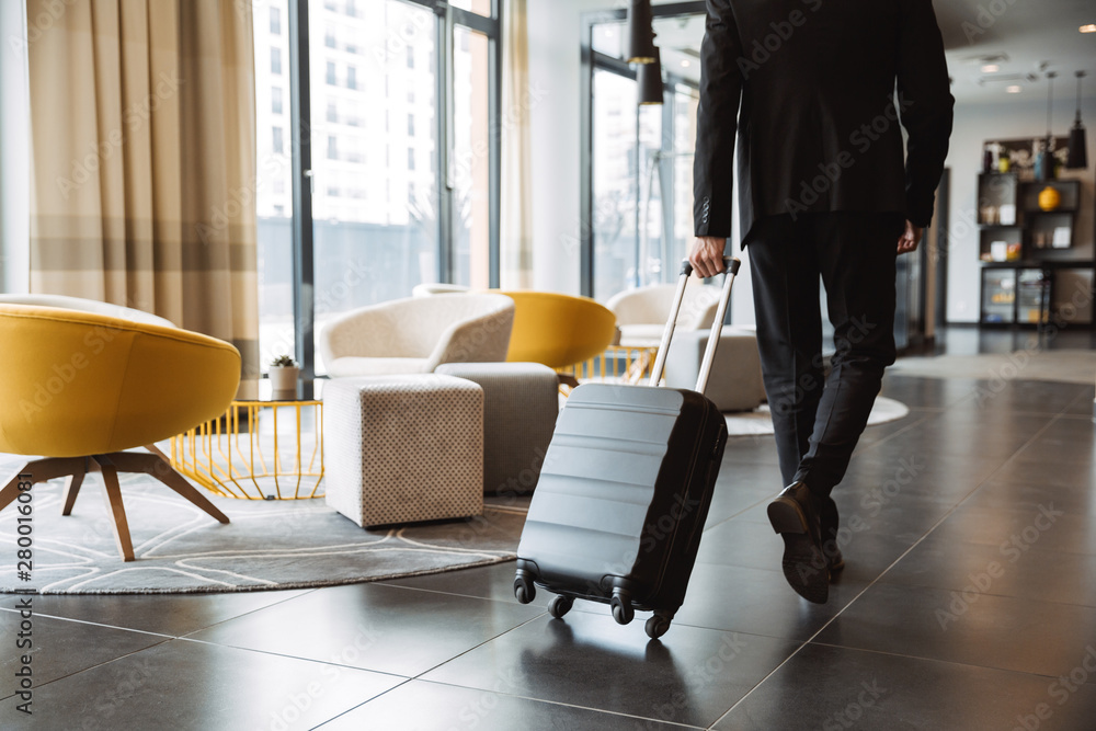 Cropped photo of caucasian businessman wearing suit walking with suitcase in hotel lobby - obrazy, fototapety, plakaty 