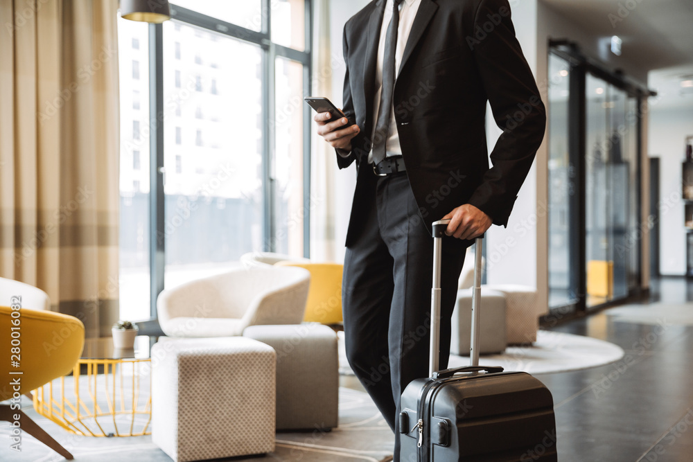 Cropped photo of confident businessman wearing suit holding smartphone and walking with suitcase in hotel lobby - obrazy, fototapety, plakaty 