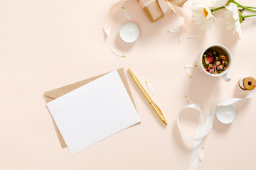 Romantic flatlay composition with blank paper card mockup, envelope, herbal tea, ribbon, daisy chamomile flower on pastel pink background. Feminine home office desk concept. Flat lay, top view.