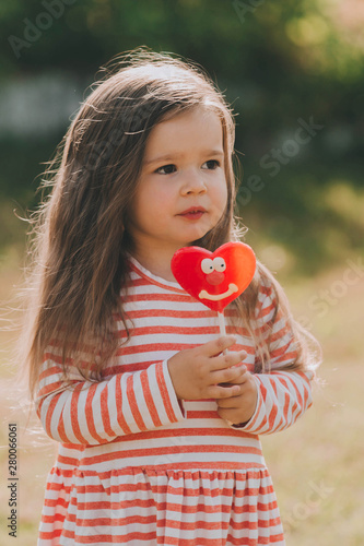 Cute Baby Girl In A Striped Dress With Long Hair And A Red Heart