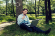 © payamona - Young and happy businessman sitting on a ground with laptop in a park and making a successful gesture Selective focus