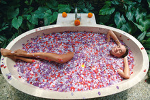 Young Woman Getting Treatment At Spa Centre. Woman Relaxing In Round Outdoor Bath With Tropical Flowers