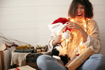 Wall Mural - Stylish happy girl in santa hat playing with cute cat in festive christmas lights on background of modern room with presents. Young hipster woman in cozy sweater hugging kitty and smiling