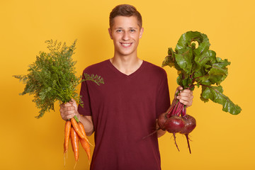 Happy cheerful young blond man holding healthy vegetables for salad. Raw foodist with carrots and beets posing isolated over yellow studio background. Raw dood diet and healthy eating concept.