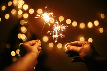 Glowing Sparklers In Hands On Background Of Golden Christmas Tree Lights, Couple Celebrating In Dark Festive Room. Happy New Year. Space For Text. Fireworks Burning In Hands. Happy Holidays
