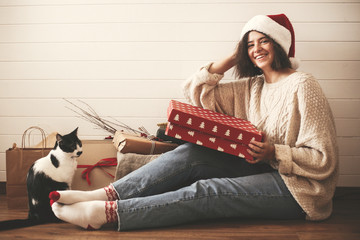 Wall Mural - Stylish happy girl in santa hat and cozy sweater playing with cat and holding christmas gift box in decorated christmas room. Young hipster woman holding present and smiling. Merry Christmas