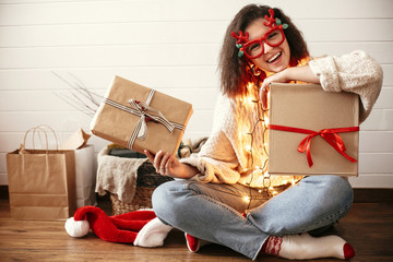 Wall Mural - Stylish happy girl in festive glasses with reindeer horns holding christmas gift boxes in christmas lights. Young hipster woman in cozy sweater holding christmas presents in modern room.