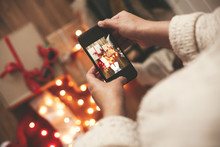 Hands Holding Phone And Taking Photo Of Christmas Gift Boxes, Santa Hat, Lights On Wooden Background In Dark Room. Stylish Hipster Girl In Sweater Making Christmas Instagram Flat Lay Photo