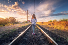 Standing Young Woman On The Railroad At Sunset In Summer. Slim Girl On The Railway Station. Rural Industrial Landscape With Railroad, Sky With Colorful Clouds. Railway Platform. Transportation. Travel