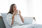 Fototapeta Psy - Young caucasian woman in white pajamas with white holding a cup of coffee and drink and find information from the laptop on a bed.