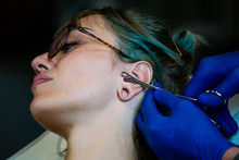 Young Woman Getting Her Ear Pierced. Man Showing A Process Of Piercing With Steril Medical Equipment And Latex Gloves. Body Piercing Procedure