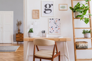 Design scandinavian interior of home office space with stylish chair, wooden desk, ladder, cube, commode, elegant accessories and mock up posters frames. Stylish home decor. Template. White wall. 