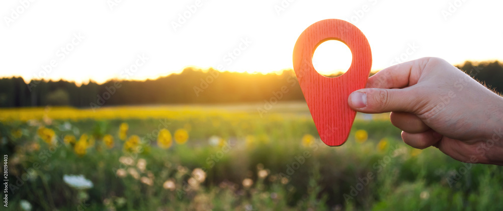 A hand is holding a red location marker in the sunset background. The concept of tourism and travel. Navigation and exploration. Destination. Holiday, vacation. Buying building land. Selective focus - obrazy, fototapety, plakaty 