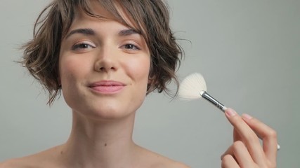 Poster - Cute young lady with natural skin making make-up using brush while looking at the camera over grey background isolated