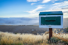 Wooden Vintage Mailbox In Beautiful Nature Landscape, Golden Dried Grass With Small Flower And Sea Of Fog And Blue Sky In The Background In Cold Weather Morning