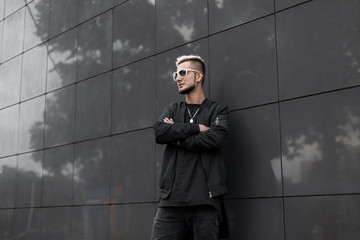 Wall Mural - American young hipster man with a fashionable hairstyle with a beard in stylish sunglasses in a trendy black jacket posing near a gray wall on the street. Handsome urban guy walks in the city.