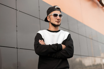 Wall Mural - Stylish young hipster man in stylish sweatshirt in black trendy black baseball cap in white glasses in the city. Handsome guy model posing near the gray wall. New youth collection of men's clothing.